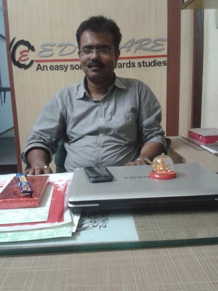 A man in a striped shirt at a desk, with the EDU-CARE sign on the wall behind him